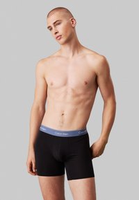 Fekete pamut boxer alsónadrág kék derékkal, amelyen a "Calvin Klein" márkaneve szerepel. A fazon szoros, mintázat nélkül.