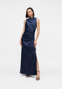 Γυναίκα που φοράει αμάνικο σατέν φόρεμα σε μπλε ναυτικό χρώμα με ψηλό λαιμόκοψη και σκίσιμο στο πλάι, συνδυασμένο με μαύρες πέδιλες σε λευκό φόντο.