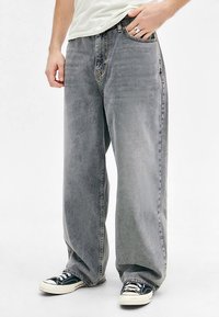 Man wearing light gray wide-leg jeans and black low-top sneakers, with one hand in the pocket and a white shirt partially visible.