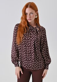 Blusa marrone con dettaglio a fiocco al collo, caratterizzata da un motivo a pois rosa chiaro. Maniche lunghe a palloncino con polsini. Texture liscia.