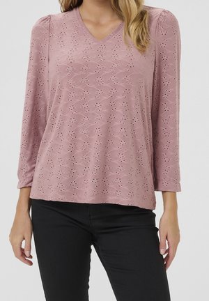 Femme portant un chemisier mauve clair à manches longues avec broderie œillets et un pantalon noir sur un fond uni.