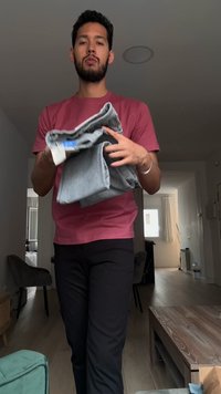 Homme en chemise rouge tenant un pantalon gris plié, debout dans un salon avec une décoration neutre et de la lumière naturelle.