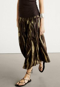 Bruine rok met een patroon van verticale gouden strepen, vloeiende vorm, lichtgewicht stof. Gecombineerd met zwarte sandalen met bandjes en een zilveren armband.