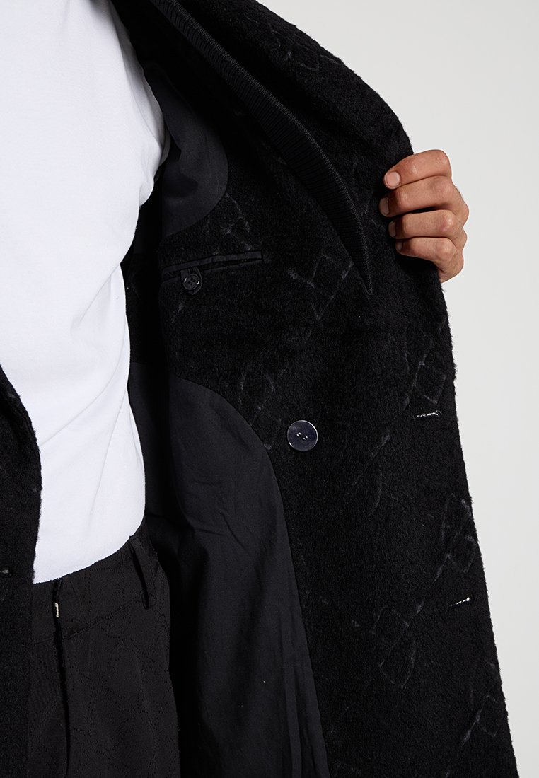 Manteau en laine noir avec une surface texturée, doté d'une fermeture à boutons et d'une doublure intérieure. L'intérieur présente un tissu noir lisse.