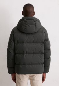 Mężczyzna w ciemnozielonej kurtce puchowej z kapturem, stojący tyłem do gładkiej jasnej ściany, pod kurtką widoczne beżowe spodnie.