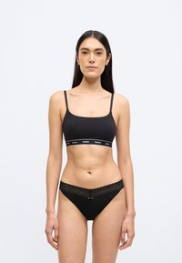 Braletă neagră cu bretele subțiri și bandă cu logo, asortată cu chiloți negri cu dantelă la margini, având un detaliu subtil cu fundiță în față.