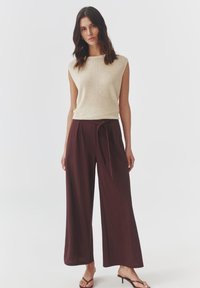 Beige bezrukávový pletený top s okrúhlym výstrihom v kombinácii s nohavicami s širokými nohavicami v burgundskej farbe, ktoré majú predné viazanie. Modelka má na sebe sandále na remienky.