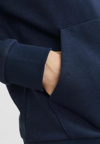 Marineblauwe sweatshirt met geribbelde mouwen, met een voorvak en een zachte textuur. Close-up toont een hand in de zak, waardoor de ontwerpinformatie wordt benadrukt.