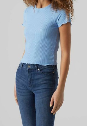 Frau trägt ein eng anliegendes hellblaues geripptes Kurzarmshirt und hoch taillierte dunkelblaue Jeans, steht vor einem einfarbigen grauen Hintergrund.