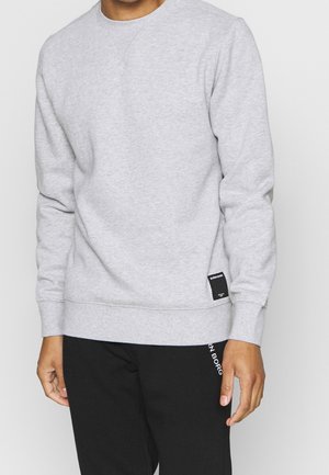 Mand iført lysegrå langærmet sweatshirt og sorte bukser, stående med armene afslappet ned langs siderne mod en ensfarvet baggrund.