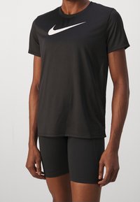 Camiseta atlética negra hecha de una tela suave y ligera, con un prominente logo blanco de Nike en el pecho. Modelo vistiendo pantalones cortos negros.