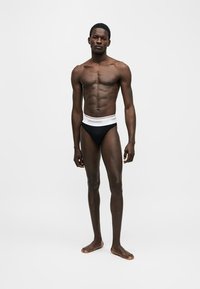 Άνδρας μοντέλο στέκεται ξυπόλητος, φορώντας μαύρα σλιπ με λευκή μέση, σε απλό λευκό φόντο.