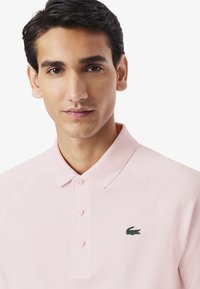 Jasnoróżowa koszulka polo z teksturowanej tkaniny, z ribbowanym kołnierzykiem, trzema guzikami oraz małym zielonym logo krokodyla na piersi.