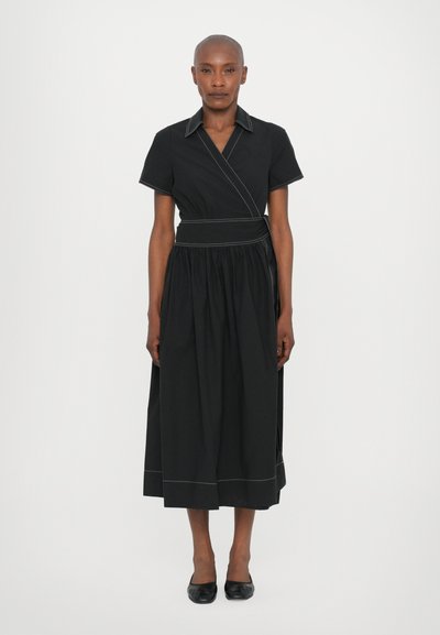 Zwarte wikkeldress met korte mouwen, kraag en een gestrikt middel. Heeft contrasterende witte stiksels en een vloeiende rok. G gedragen met zwarte schoenen.
