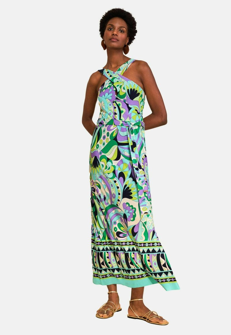 Vrouw draagt een mouwloze maxi-jurk met gekruiste bandjes en groene, paarse en blauwe abstracte bloemmotieven en gouden sandalen met bandjes.