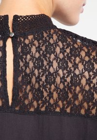 Blusa negra con un yugo de encaje con patrones florales, cierre de botones en la parte posterior y una tela suave debajo de la sección de encaje.