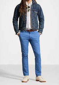 Person som bär en jeansjacka över en vit skjorta, blå byxor med ett brunt flätat bälte, beige klänningsskor och solglasögon fastklippta på skjortan.