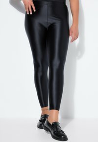 Zwarte leggings met hoge taille, gemaakt van een glad, glanzend materiaal en een slanke pasvorm. Draagt zwarte loafers met franjes als detail.