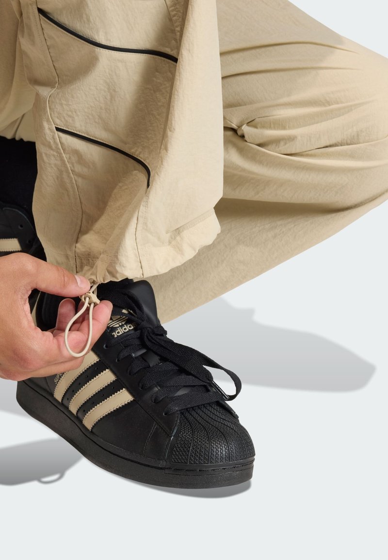 Sneakers nere Adidas con punta in gomma strutturata, abbinate a pantaloni beige. I pantaloni presentano cuciture nere e un cordoncino alla caviglia.