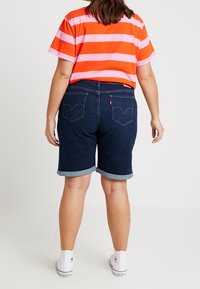 Shorts de mezclilla azul oscuro con puños enrollados, que cuentan con bolsillos traseros con un logotipo de la marca y se combinan con una camiseta a rayas naranja y rosa.