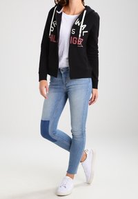 Svart zip-up hoodie med röd och vit text, parat med ljusblå jeans som har en mörkblå lapp; vita sneakers kompletterar outfiten.