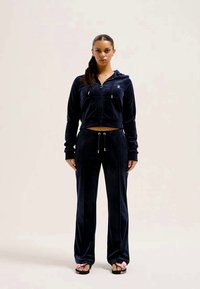 Fato de treino em veludo navy, apresentando uma hoodie curta com fecho e calças combinando com cintura elástica. O design inclui discretos detalhes de logótipos.