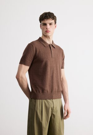 Mladić s kratkom kovrčavom kosom, odjeven u smeđu polo majicu kratkih rukava od pletenine i maslinasto zelene hlače visokog struka, stoji bočno okrenut.