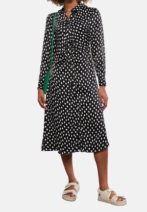 Robe noire à boutons avec des pois blancs, manches longues, taille cintrée et un ourlet au genou, associée à des sandales de couleur claire.