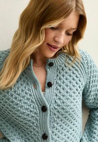 Jasnoniebieski sweter z dzianiny z dużymi brązowymi guzikami, teksturowanym wzorem, prążkowanym kołnierzem i małym wycięciem w pobliżu guzików.