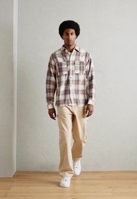 Plaid flannel shirt in beige, rust, and blue tones with two chest pockets, paired with beige trousers and white sneakers on a wooden floor.