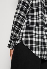 Camicia a scacchi bianca e nera con maniche lunghe, polsini con bottone e orlo curvo. Tessuto morbido con una finitura texturizzata, abbinata a pantaloni scuri.