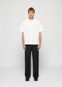 Young man standing, wearing plain white oversized T-shirt, black trousers, black shoes, and a gold pendant necklace, neutral gray background.