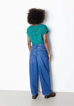 Personne aux cheveux bouclés se tient face à un mur blanc, portant un haut bleu sarcelle à dos en dentelle, un jean large bleu et des chaussures marron.