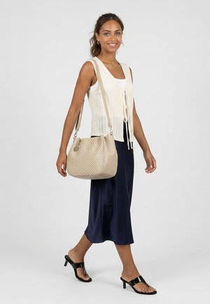 Frau lächelnd, gehend; trägt weißes Strickweste, dunkelblauen Rock, schwarze Sandaletten mit Absatz und trägt eine beige Umhängetasche.