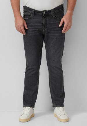 Man die een zwarte slim-fit jeans, witte sneakers en een wit ingestopt shirt draagt, staand tegen een effen lichtgrijze achtergrond.