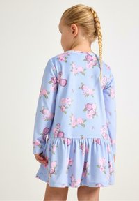 Dziecko z długimi zaplecionymi warkoczami, ubrane w jasnoniebieską sukienkę ozdobioną różowymi i fioletowymi kwiatowymi wzorami, zwrócone tyłem na jednolitym tle.