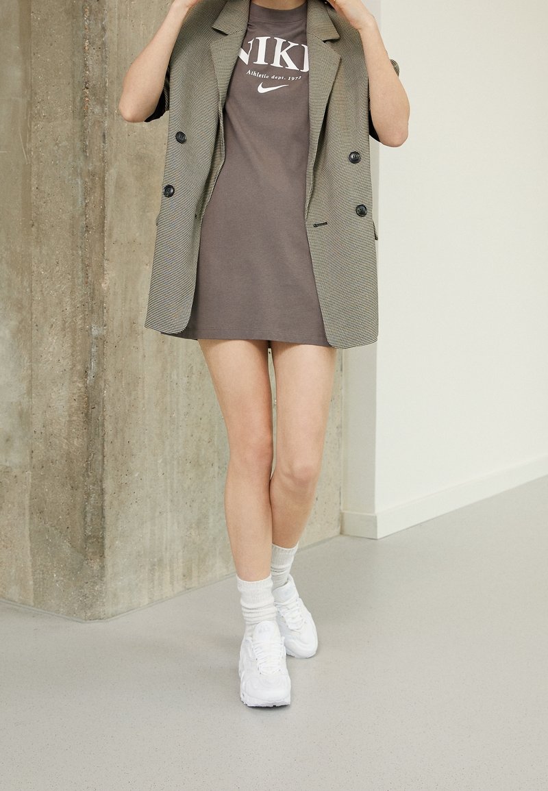 Blazer grigio a scacchi indossato sopra un vestito a t-shirt con grafiche marroni della Nike, abbinato a sneakers bianche e calzini a righe, su un pavimento liscio.