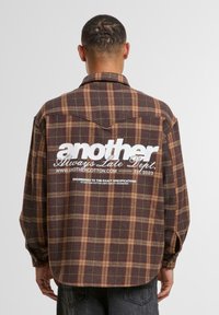 Brown and tan plaid flannel shirt with a curved hem, buttoned cuffs, and large white text logo on the back.