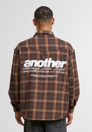 Brown and tan plaid flannel shirt with a curved hem, buttoned cuffs, and large white text logo on the back.