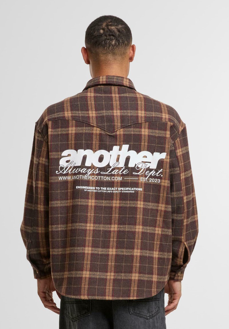 Brown and tan plaid flannel shirt with a curved hem, buttoned cuffs, and large white text logo on the back.