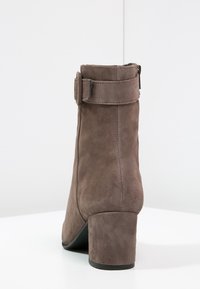 Bottine en daim marron avec un talon carré, dotée d'une sangle et d'une boucle sur le dessus. Texture lisse et design minimaliste.
