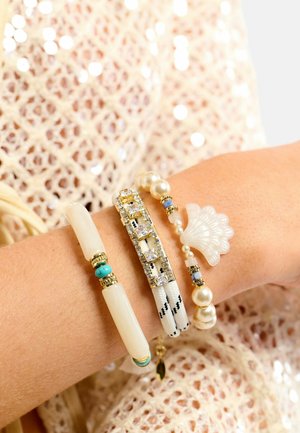 Quatre bracelets sur un poignet : un perle en forme de corne blanche avec des accents turquoise, un coquillage avec des accents dorés, un bracelet extensible orné de strass et des perles de culture.