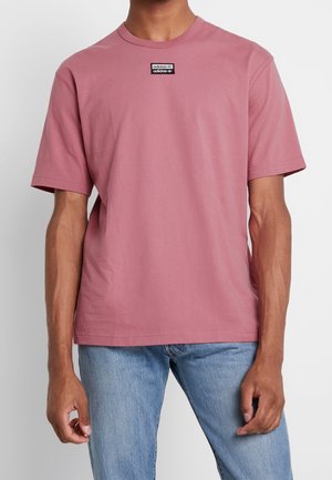 Homme portant un t-shirt Adidas rose poudré uni avec un petit patch logo noir et blanc sur la poitrine, associé à un jean bleu clair.