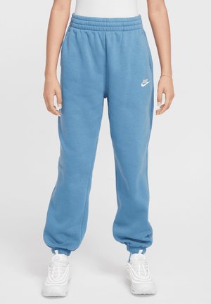 Person iført lyseblå Nike joggingbukser med elastiske manchetter og hvide sneakers, stående mod en ensfarvet baggrund.