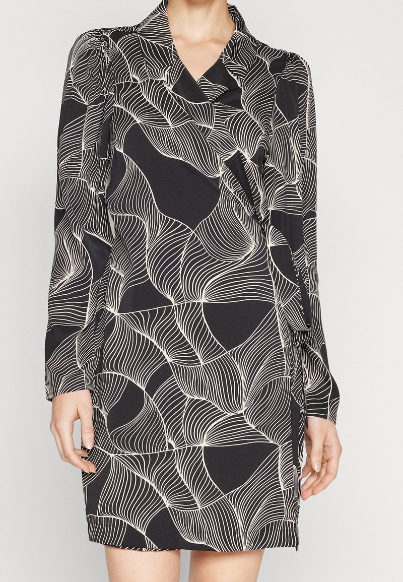 Robe noire à manches longues avec un motif abstrait de lignes ondulées blanches et un large col, portée par une personne debout sur un fond uni.
