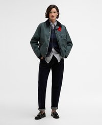 Casaco verde, impermeável, com um colarinho escuro e detalhes em vermelho, sobreposto a um suéter de malha azul-marinho e uma camisa riscada, combinado com calças de ganga escuras.