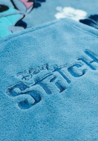 Coperta in pile blu con testo ricamato "Disney Stitch". La texture è morbida, con una finitura liscia e una leggera lucentezza.