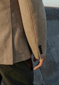 Blazer beige texturizado con un primer plano de la manga, que cuenta con cinco botones negros. La tela tiene un aspecto tejido y un corte estructurado.