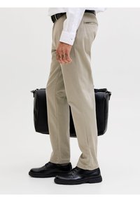 Άνδρας που φοράει μπεζ παντελόνι, λευκό πουκάμισο, μαύρα παπούτσια, κρατώντας μια μαύρη δερμάτινη τσάντα μπροστά σε απλό λευκό φόντο.