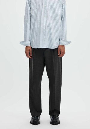 Persona che indossa una camicia a righe azzurro chiaro con bottoni, pantaloni larghi grigio scuro e scarpe nere testurizzate, in piedi davanti a uno sfondo semplice.